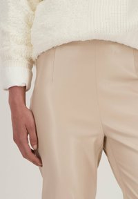 Beige neplederen broek met een gladde afwerking, gecombineerd met een witte trui met textuur, die mouw.details onthult.