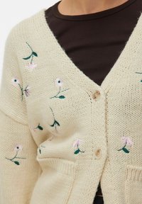 Kremasta pletena jopica z rožnim vezom v roza in zeleni barvi, z V-izrezom in enojno zaponko. Mehka struktura in sproščen kroj.