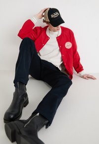 Rote Windbreaker-Jacke mit weißen Akzenten und Logo, cremefarbenes T-Shirt, marineblaue Hose und schwarze Leder-Stiefeletten, sitzend vor einem schlichten Hintergrund.