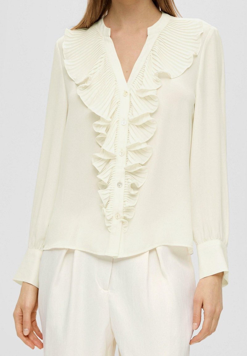 Blusa bianca con fronte arricciato, maniche lunghe, chiusura con bottoni e un tessuto morbido e leggero. Presenta un colletto e polsini texturizzati.