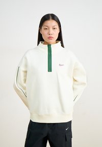 Kremai krāsas treniņjakas ar augstu apkakli, ceturtdaļgaruma zaļu rāvējslēdzēju un mazu rozā logotipu. Piedurknēm ir zaļi akcenti.