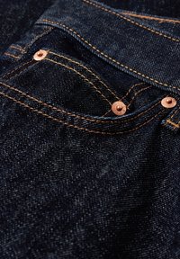 Närbild av en ficka på mörkblå denimjeans med kopparnitar och orange sömnad.