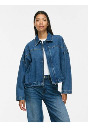Kvinde iført en løs blå denimjakke over en hvid skjorte og matchende denimjeans, stående foran en ensfarvet hvid baggrund.
