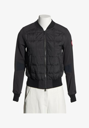 Veste bomber noire matelassée avec fermeture éclair à l'avant, poignets et taille côtelés, portée sur un mannequin avec un pantalon blanc.