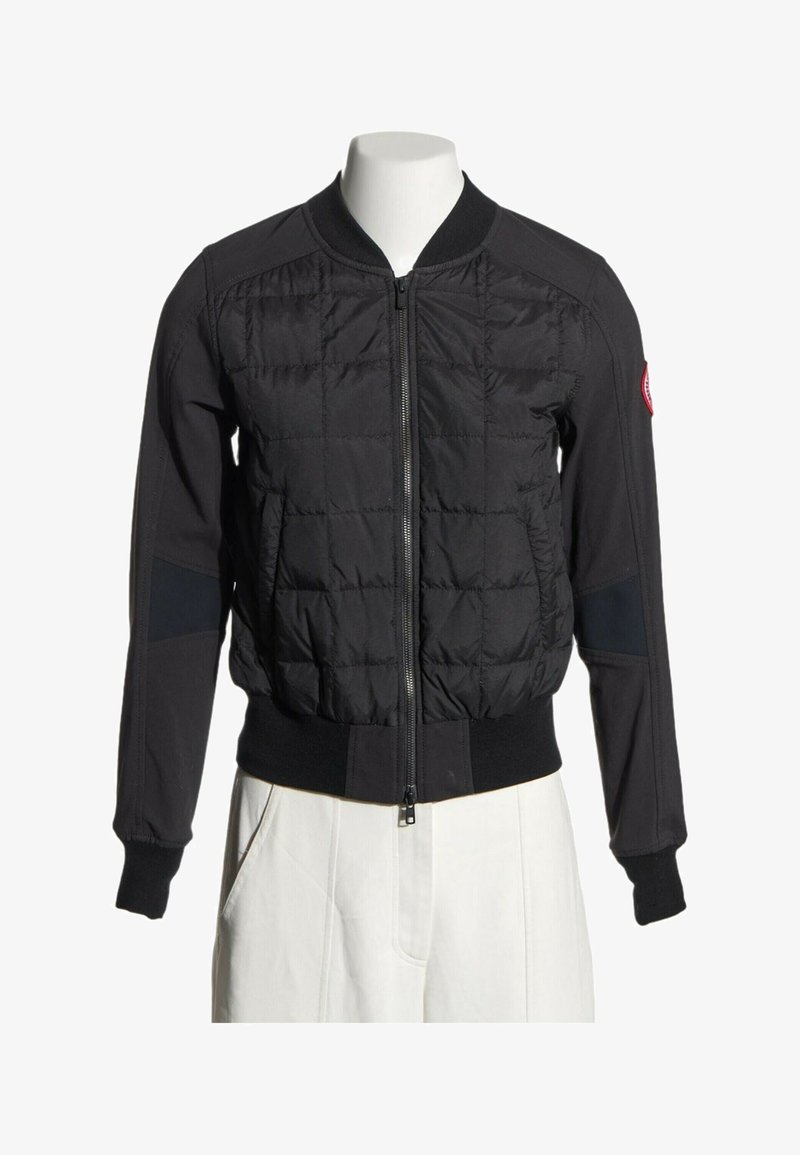 Veste bomber noire matelassée avec fermeture éclair à l'avant, poignets et taille côtelés, portée sur un mannequin avec un pantalon blanc.