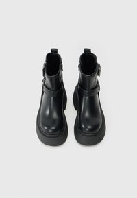 Botas de tobillo de cuero negro con punta redonda, textura suave, suela de plataforma gruesa, cremallera lateral y detalle decorativo de correa cerca del tobillo.