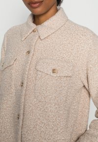 Giacca beige texturizzata con colletto, due tasche sul petto e chiusura con bottoni. Il tessuto presenta un aspetto morbido e peloso.