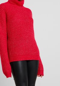 En kraftig röd polotröja i texturerad stickad kvalitet, med oversize ärmar och ribbat nederkant, i kombination med svarta läderbyxor.