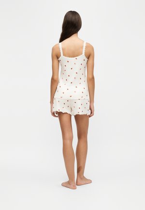 Femme aux longs cheveux foncés debout pieds nus, vêtue d'un haut blanc à fines bretelles et d'un short avec un petit motif de fruits orange-rouge, tournée de dos.