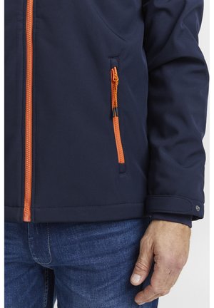 Veste marine avec des fermetures éclair orange, comprenant une poche poitrine. Présente une texture lisse, des poignets élastiques et une coupe ajustée.