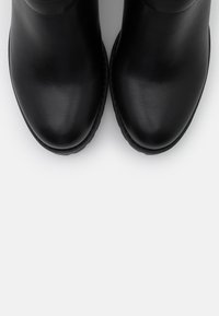 Bottes basses en cuir noir à texture lisse, bout arrondi, avec un détail de couture subtil le long de la base. Design minimaliste, sans quincaillerie visible.