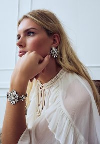 Sparkling silver statement earrings with leaf-like shapes, paired with an ornate bracelet of similar design, worn with a white ruffled blouse.