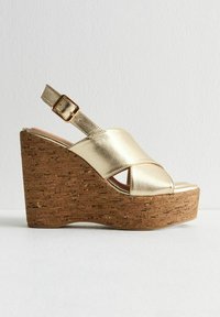 Sandale à talon en métal doré avec une base en liège. Présente un design à bout ouvert et une sangle réglable à boucle. Texture lisse sur l'ensemble.