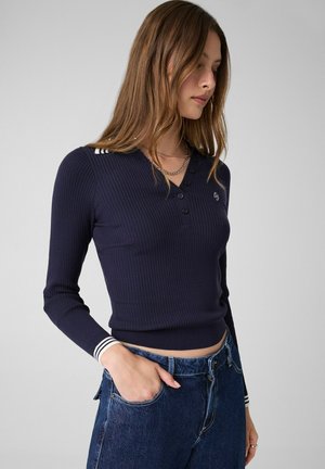 Ung kvinde med langt hår iført en marineblå ribbet langærmet poloshirt og blå jeans med den ene hånd i lommen mod en ensfarvet baggrund.