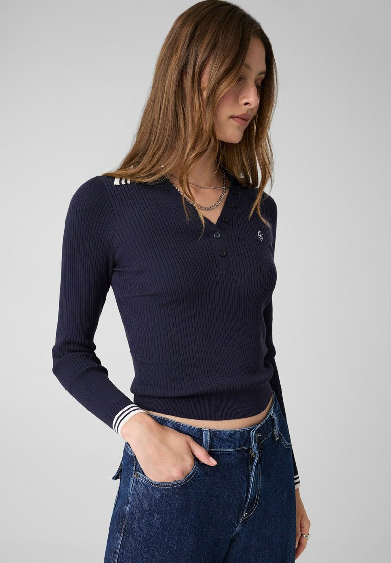 Jeune femme aux longs cheveux, portant un polo bleu marine côtelé à manches longues et un jean bleu, une main dans la poche, sur un fond uni.