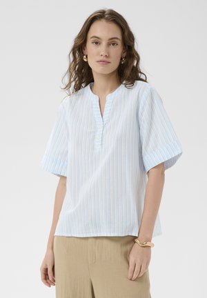 Donna con capelli mossi che indossa orecchini a cerchio dorati, una blusa a righe bianche e azzurre a maniche corte, pantaloni beige e un braccialetto d'oro.