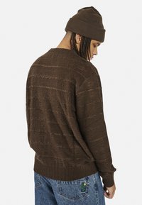 Maglione marrone lavorato a maglia con motivi testurizzati, dotato di orlo e polsini a coste. Indossato con jeans in denim blu, con una toppa a forma di cactus.