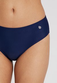 Marineblauwe bikini onderkant met een middelhoog snit, een gladde textuur en een zilveren logo-accent aan de zijkant. Strakke lijnen en een minimalistisch ontwerp.