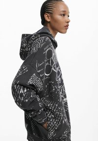 Zwarte hoodie met witte handgeschreven tekst en illustraties, met een losse pasvorm, lange mouwen en een koord in de capuchon.