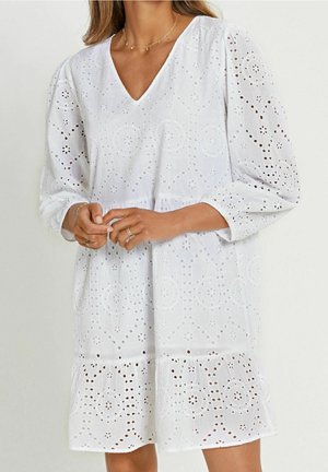 Femme portant une robe blanche en dentelle eyelet avec un col en V, des manches longues bouffantes et un ourlet volanté, accessoirisée de bagues en or et d’un collier délicat.