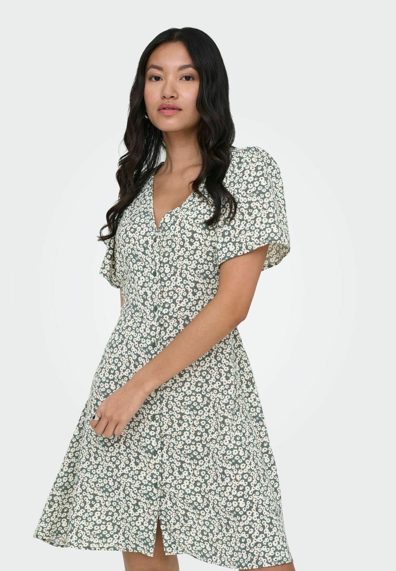 Femme aux cheveux longs et foncés, portant une robe verte à manches courtes avec un imprimé floral blanc, debout devant un fond uni.