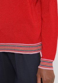 Roter Strickpullover mit glitzender Textur; mit gestreiften Rippbündchen und Saum in Pink, Schwarz und Rot. Halb lange Ärmel, taillierte Passform.