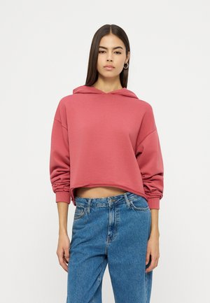 Kobieta ubrana w różowy, krótki, zgaszony różowy sweter z długimi rękawami i niebieskie, wysokie dżinsy, stojąca na białym tle.