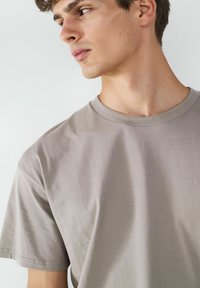 Jasnoszara koszulka z okrągłym dekoltem, wykonana z miękkiej bawełny. Krótkie rękawy, luźny krój, gładka tekstura z subtelnymi detalami szwów na brzegach.