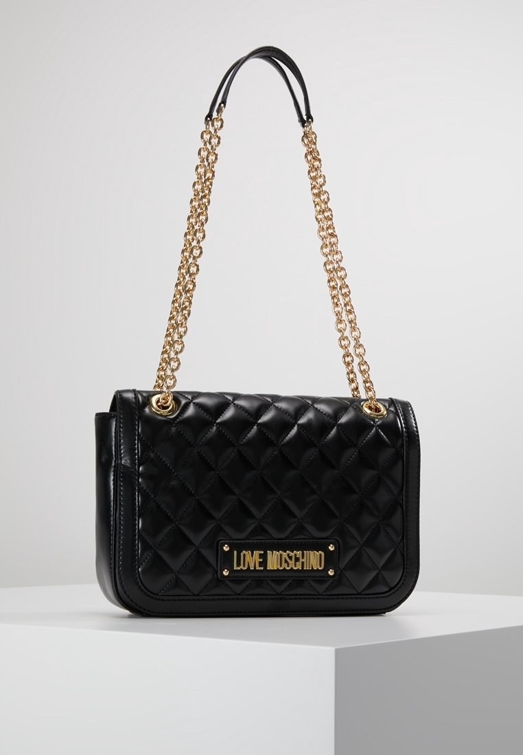 Svart vadderad handväska i konstläder med ett guldkedjespänne och metall-logoplåt. Har en strukturerad rektangulär form och en slät yta.