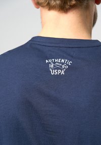 Navyblaues T-Shirt mit kurzen Ärmeln, das auf dem oberen Rücken mit einem weißen Logo-Text versehen ist, der "AUTHENTIC 1890 USPA" liest. Glattes Baumwollgewebe.