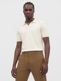 Mand med fletninger iført en cremefarvet poloshirt med korte ærmer og brune bukser, stående mod en ensfarvet lys baggrund.