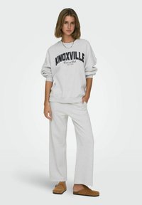 Sivi, preveliki dukserica s natpisom "KNOXVILLE" u crnom textu, u kombinaciji s odgovarajućim sivim širokim hlačama i smeškim klompama.