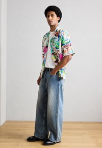 Άνδρας που στέκεται με τα χέρια στις τσέπες, φοράει πολύχρωμο πουκάμισο με σύντομο μανίκι και σχέδια, λευκή μπλούζα, φαρδιά μπλε τζιν και μαύρα παπούτσια πάνω σε ξύλινο πάτωμα.