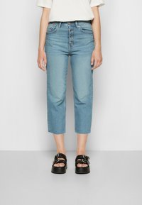Jeans de ganga azul claro com cintura alta, frente de cinco botões, comprimento cropped e bainhas com acabamentos crus, combinados com sandálias de plataforma pretas e grossas.