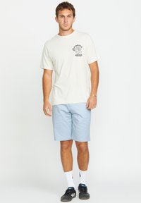 Light beige short-sleeve t-shirt with black graphic on chest, paired with light blue shorts. Model wearing black sneakers and white socks.
