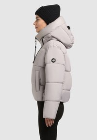 Giacca puffer grigia chiara con cappuccio, caratterizzata da una tasca frontale, una toppa con logo sul braccio e una texture liscia. Indossatore con pantaloni neri e un berretto.