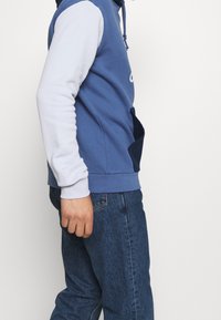 Hooded sweatshirt with a blue body, white sleeves, and a large front pocket. Soft fabric with a smooth finish and visible stitching.