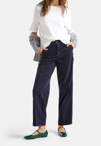 Kvinna iklädd vit t-shirt, marinblå kordfluwelepants, grå cardigan hängande över armarna och blanka gröna Mary Jane-skor, stående med händerna i fickorna.