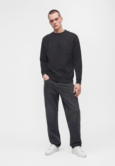 Jeune homme portant un sweat-shirt noir avec le texte "ICEBERG since 1971", un jean foncé et des baskets blanches, debout devant un fond blanc uni.