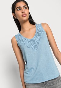 Top sem mangas azul claro com um decote decorativo em renda com padrões intrincados. O tecido parece suave com uma textura lisa.