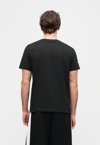 Άνδρας που φαίνεται από πίσω, φοράει απλή μαύρη κοντομάνικη μπλούζα και μαύρο σορτς με λευκές κάθετες ρίγες στα πλαϊνά.