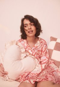 Rózsaszín mintás hálóruha fodros szegélyekkel, szív alakú csíkos párnát tartva. A háttérben lágy textúrák és színek dominálnak.