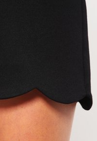 Jupe en tissu noir avec un ourlet festonné. La texture semble lisse, avec un léger éclat. La jupe est ajustée et ne présente aucun motif visible.