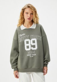 Olijfgroene sweatshirt met een witte kraag, met daarop bedrukhte graphics: "Florida WEST COAST 89 MIAMI SUNSET BEACH EST. 1993." Oversized pasvorm.