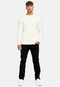 Weißer, kabelgestrickter Pullover mit strukturierten Mustern, kombiniert mit schwarzen Hosen und weißen Sneakers, Modell posiert vor einem schlichten Hintergrund.