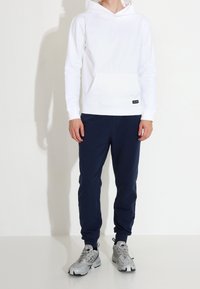 Felpa bianca con cappuccio e tasca frontale, abbinata a pantaloni della tuta blu navy e scarpe da ginnastica grigie. Tessuto morbido, vestibilità rilassata e design minimalista.