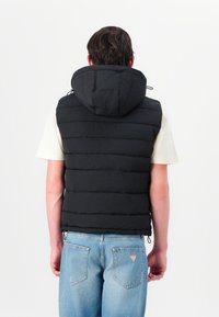 Vesta puffer neagră cu textură matlasată orizontală, glugă și șnururi ajustabile. Pur wearable peste o cămașă de culoare deschisă, asortată cu blugi albaștri.