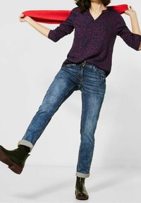 Mujer con una blusa azul de estampado y jeans arremangados, con botas negras, sosteniendo una bufanda roja detrás de su cuello.