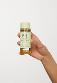 Klar glasflaske med Pixi Vitamin C Tonic, cylindrisk form, lys grøn kapsel. Væsken er amberfarvet; etiketten har grøn og gul tekst.