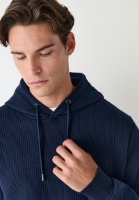 Laivastonsininen ribbineulosta oleva huppari, jossa on kiristysnauhallinen huppu ja hopeiset metalliset nauhanpäät; siinä on pitkät hihat ja teksturoitu kangas.